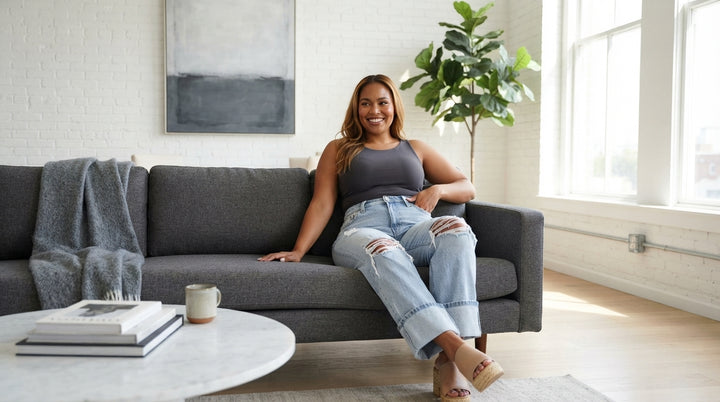 A woman wearing distressed high-waist jeans relaxing on a charcoal sofa in a bright, modern living room.